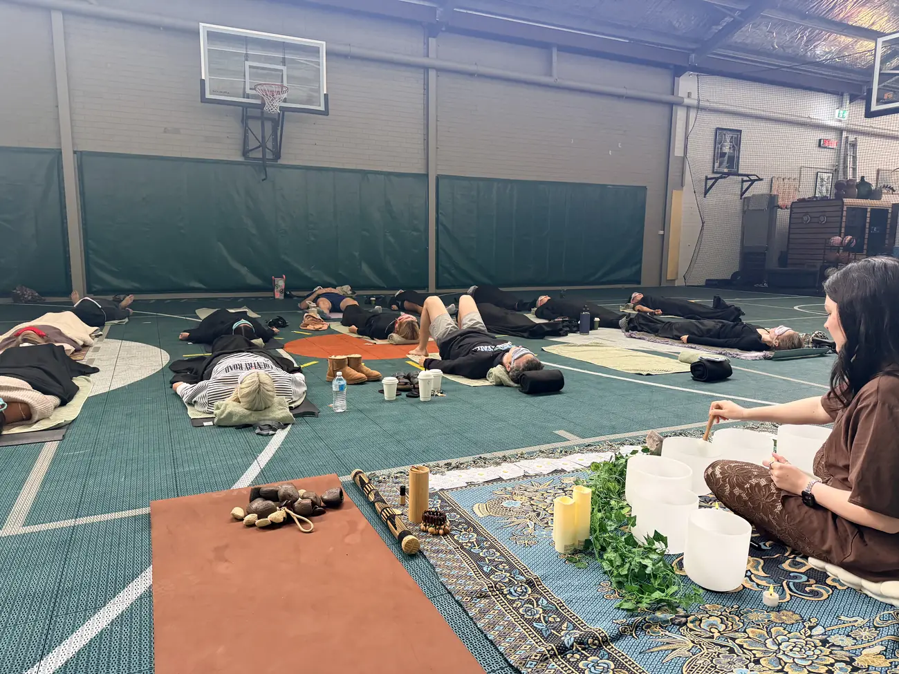 Steph playing crystal singing bowls for a group resting on mats on the indoor court at Ark Sports Club, Burswood
