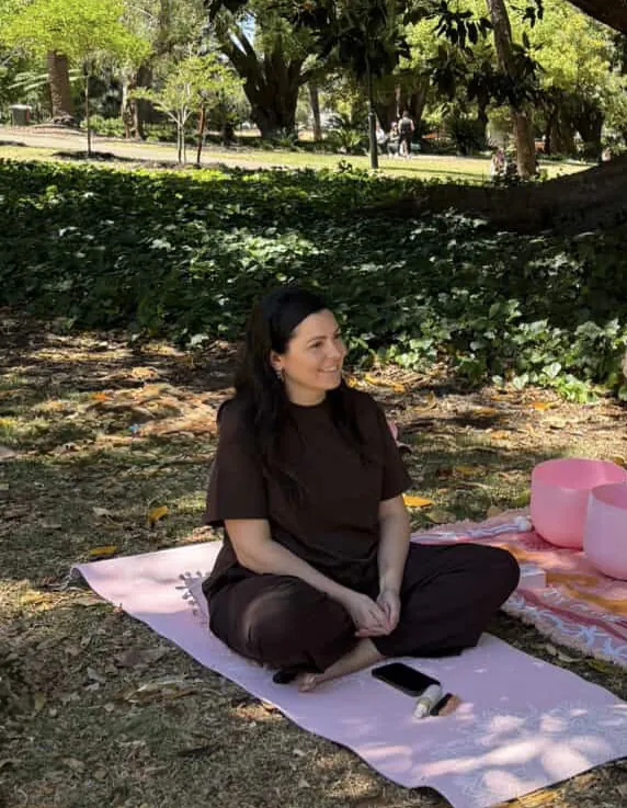 Steph sitting on a mat with crystal singing bowls outdoors under the trees